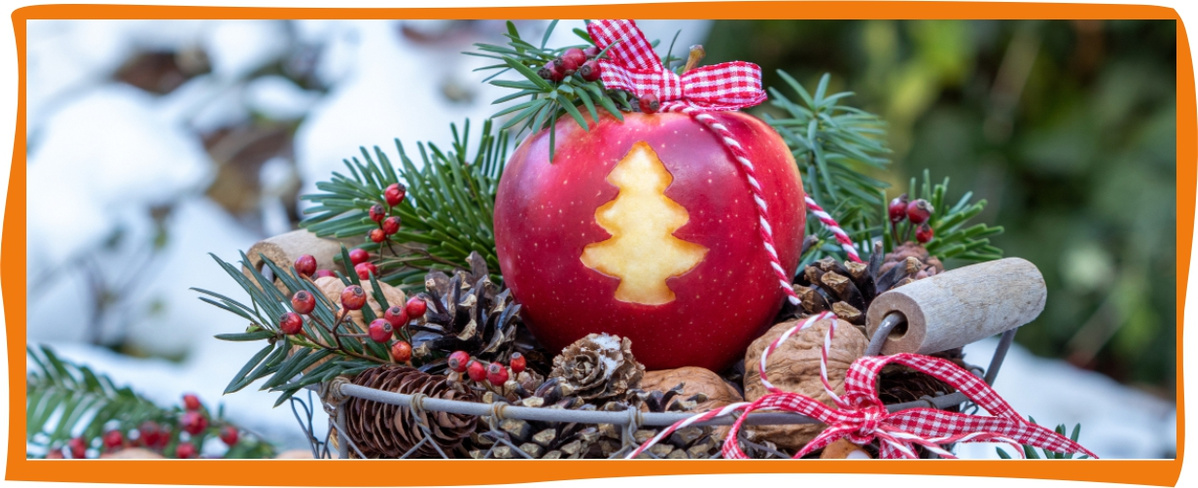 KI generiert: Das Bild zeigt einen dekorativen Korb mit einem Apfel, der eine Tannenbaumform eingeritzt hat. Der Korb ist mit Tannenzweigen, Zapfen und roten Beeren verziert und wird von einer winterlichen Kulisse umgeben.