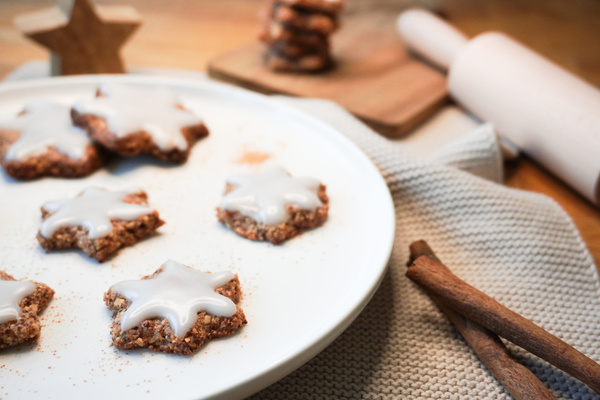 KI generiert: Das Bild zeigt sternförmige Lebkuchen mit weißem Zuckerguss auf einem Teller. Im Hintergrund sind Zimtstangen, ein Nudelholz und ein Holzbrett sichtbar, was auf eine Weihnachtsbäckerei-Szene hinweist.