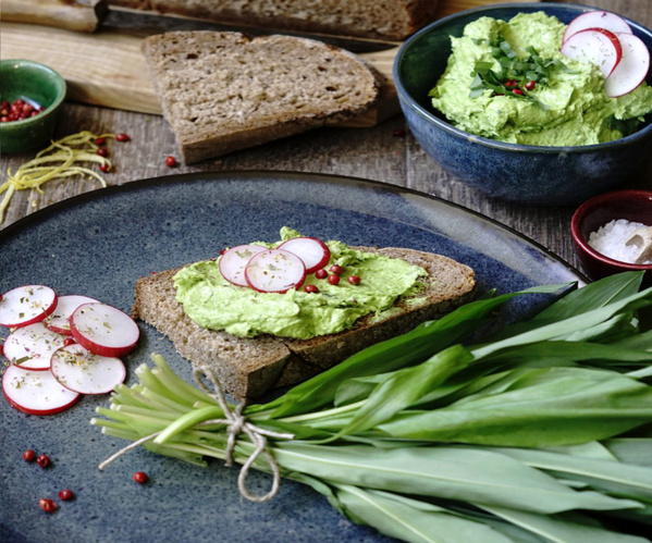 KI generiert: Brot mit grünem Aufstrich und Radieschen, umgeben von frischen Zutaten auf einem Tisch.