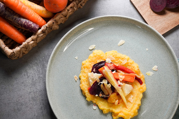 KI generiert: Das Bild zeigt ein Gericht mit cremigem Brei, garniert mit buntem, geröstetem Gemüse und Mandelsplittern. Daneben sind frische Karotten in einem Korb sichtbar.