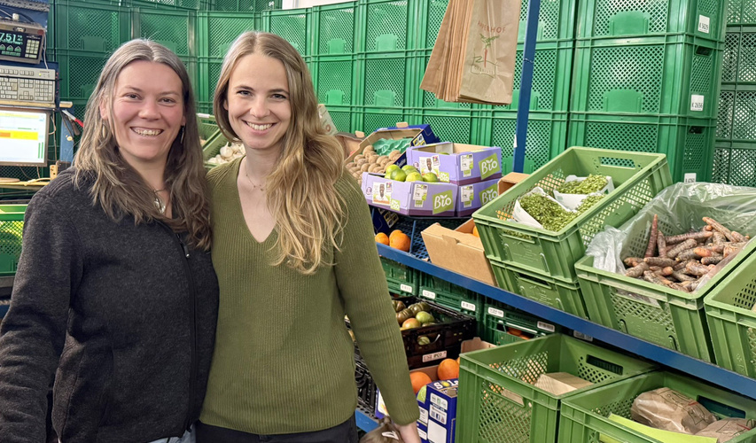 KI generiert: Zwei Personen stehen lächelnd in einem Laden mit grünen Kisten voller Obst und Gemüse.