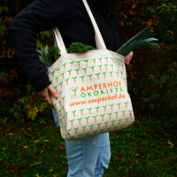 KI generiert: Die Hauptinhalt des Bildes ist eine Person, die eine bedruckte Stofftasche mit der Aufschrift "Amperhof Ökokiste" trägt. Die Tasche ist mit frischem Gemüse gefüllt.