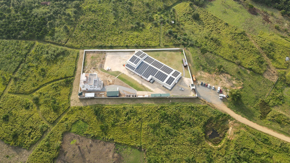 KI generiert: Das Bild zeigt eine Anlage, die von oben aufgenommen wurde und von einem Zaun umgeben ist. Auf der Anlage befinden sich Gebäude mit Solarpanelen auf den Dächern, eingebettet in eine größtenteils grüne Landschaft.