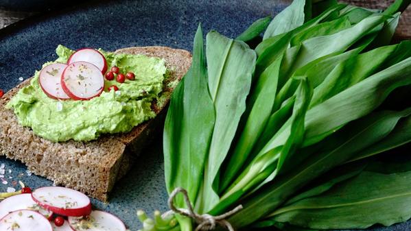 KI generiert: Ein Brot mit grünem Aufstrich und Radieschen, daneben ein Bund grüner Blätter auf einem dunklen Teller.