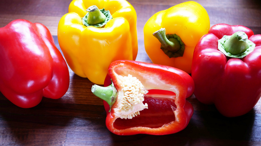 KI generiert: Rote und gelbe Paprikaschoten auf einem dunklen Holzuntergrund. Eine rote Paprika ist halbiert und entkernt.