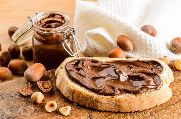KI generiert: Ein Brot mit Schokoladencreme, umgeben von Haselnüssen und einem Glas mit Creme.