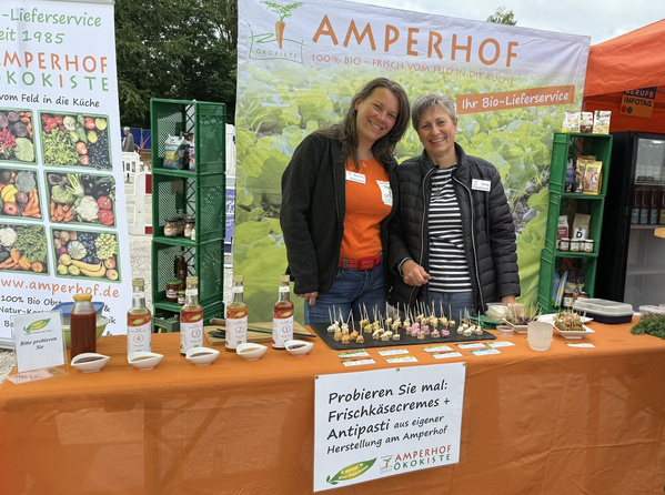 KI generiert: Zwei Personen stehen an einem Stand mit Bio-Lebensmitteln und Proben. Text: "Probieren Sie mal: Frischkäsecremes + Antipasti...".