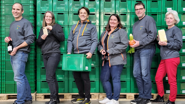 KI generiert: Eine Gruppe von sechs Menschen posiert vor grünen Kisten, einige halten Lebensmittel oder Getränkeflaschen.