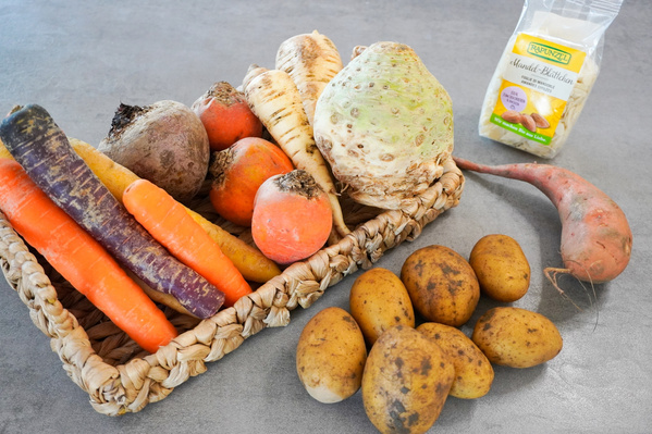 KI generiert: Das Bild zeigt eine Auswahl an frischem Wurzelgemüse, darunter Karotten, Rote Bete, Sellerie, Pastinaken und Kartoffeln sowie eine Packung Mandelblättchen. Die Zutaten liegen auf einer grauen Oberfläche und sind größtenteils in einem geflochtenen Korb arrangiert.