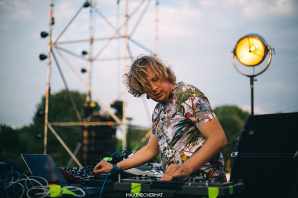 KI generiert: Ein DJ legt im Freien auf; er trägt ein buntes Hemd. Text: "MAXIMECHERMAT.COM".