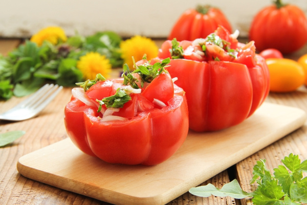 KI generiert: Gefüllte Tomaten mit Gemüse und Kräutern auf einem Holzbrett, mit buntem Gemüse im Hintergrund.