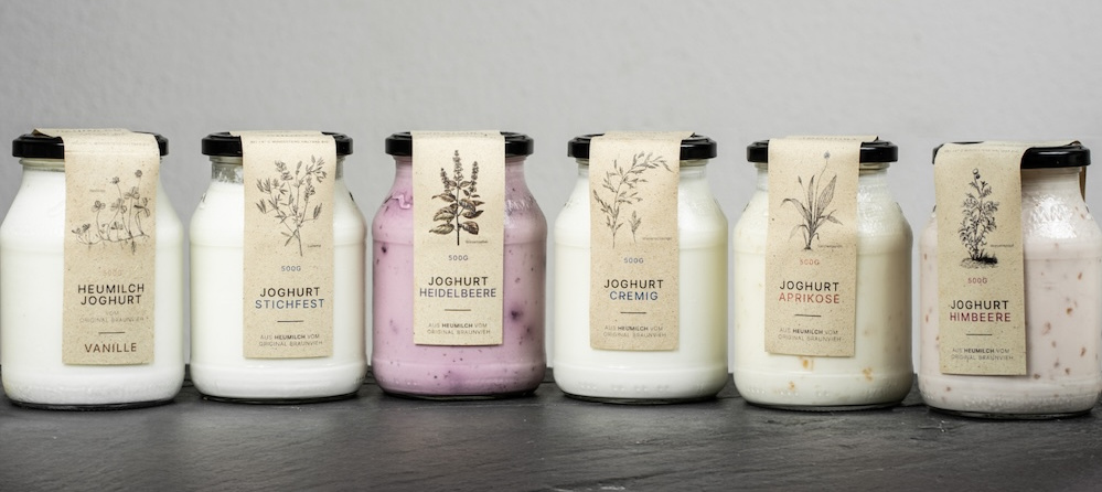 KI generiert: Sechs Gläser Joghurt mit verschiedenen Geschmacksrichtungen vor grauem Hintergrund. Text: "Heumilch Joghurt Vanille", "Joghurt Stichfest", "Joghurt Heidelbeere", "Joghurt Cremig", "Joghurt Aprikose", "Joghurt Himbeere".